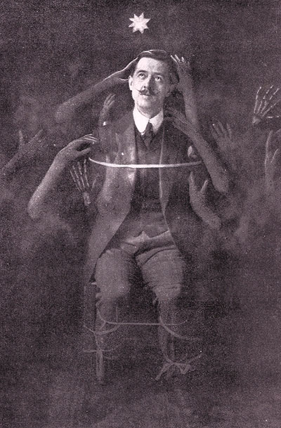A monochrome photo of a man tied to a chair, dressed in a suit. Disembodied hands are touching him and reaching for him.