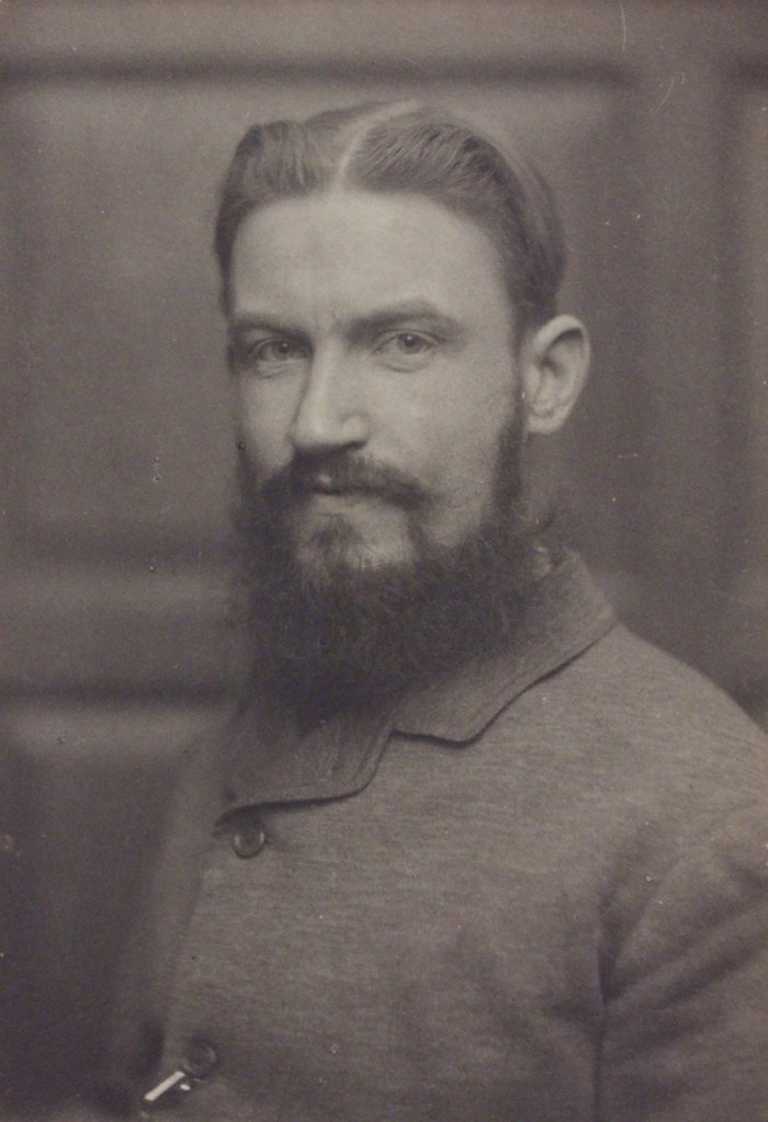 A black and white photo of the writer George Bernard Shaw. He is a white man with dark hair, swept back and centre parted. He has a moustache and a long beard. he is wearing buttoned-up jacket.