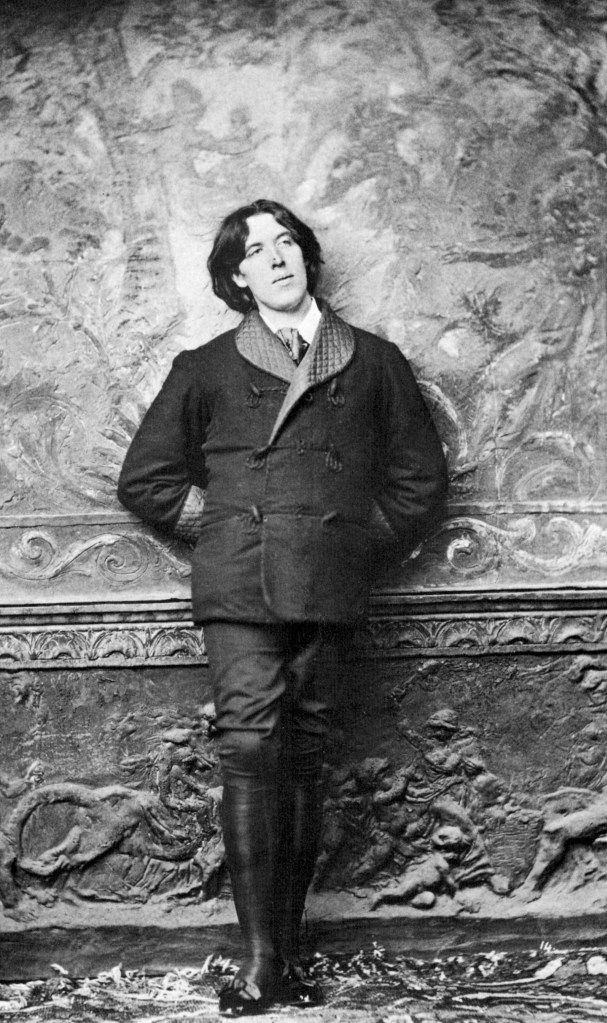 A black and white photo of the writer Oscar Wilde. He is a white man with long. dark hair parted in the middle. He is leaning against a wall wearing  a dark smoking jacket with a quilted satin collar, and dark trousers tucked into knee-length black boots.