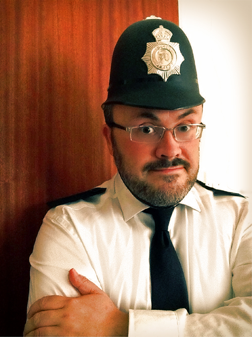 Alistair Gentry in Artbollocks Theatre: he has his arms folded and he is dressed as a police officer, except that the police helmet is much too small.