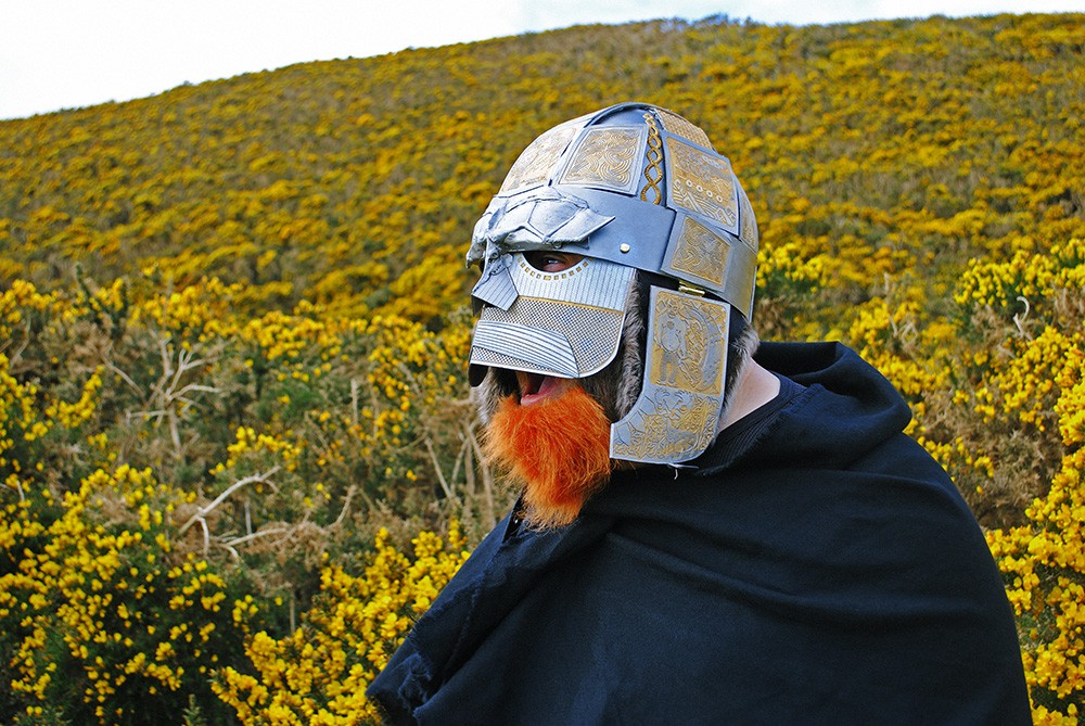 A person wearing a silver. engraved Viking helmet. They have a false ginger beard and they are shouting or screaming. In the background is a hillside covered in yellow flowering gorse.