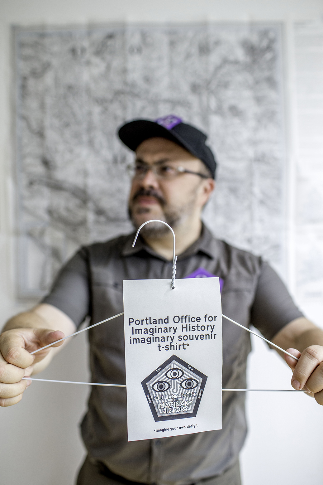 Alistair Gentry dressed as an Imaginary History ranger, holding an empty coat hanger with with a label that says "Portland Office for Imaginary History imaginary souvenir t-shirt (imagine your own design)"
