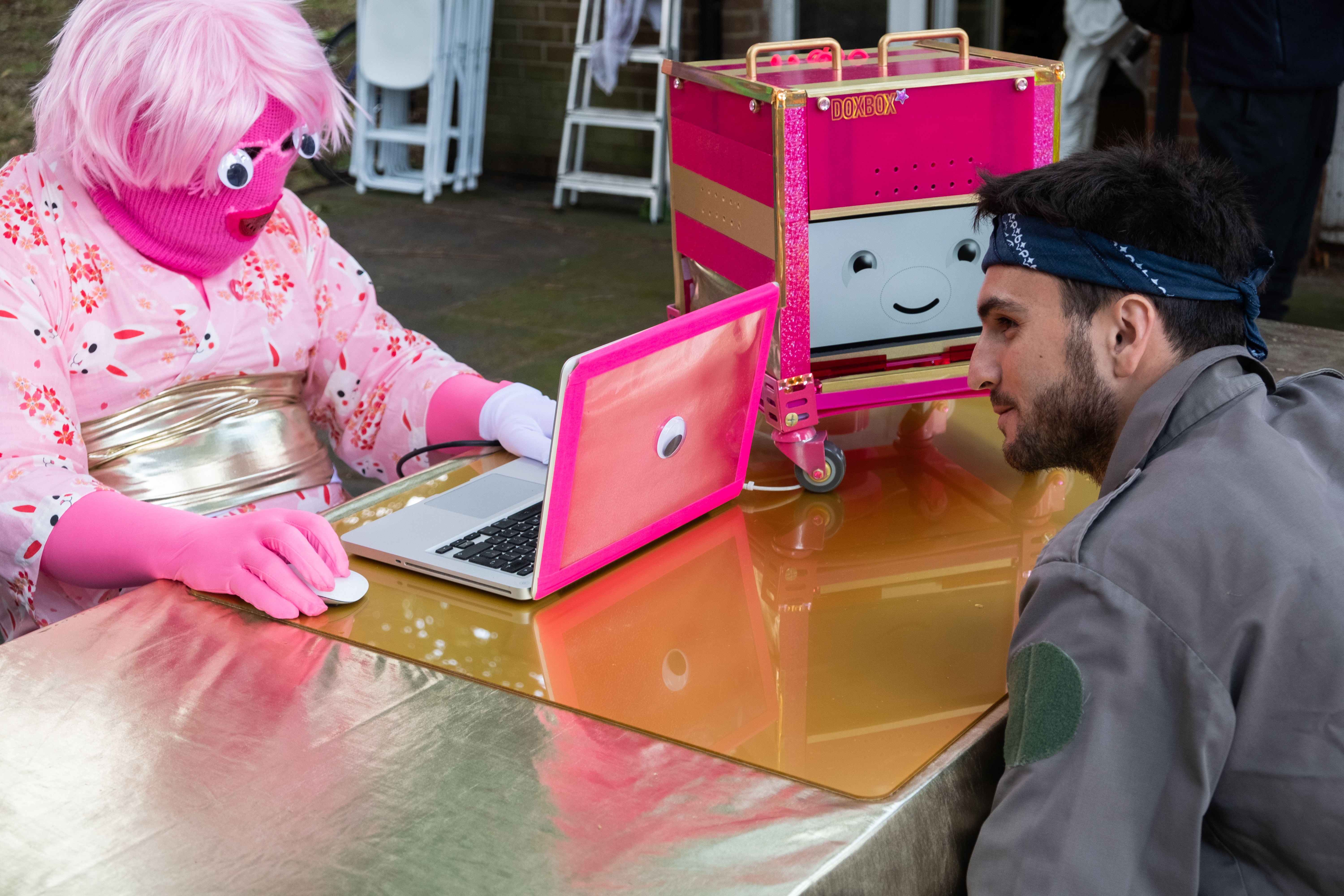 A person in a pink mask, pink wig and pink kimono, sitting at a gold desk with a pink, cube shaped robot that has a smiling face. A man is listening closely to the robot.