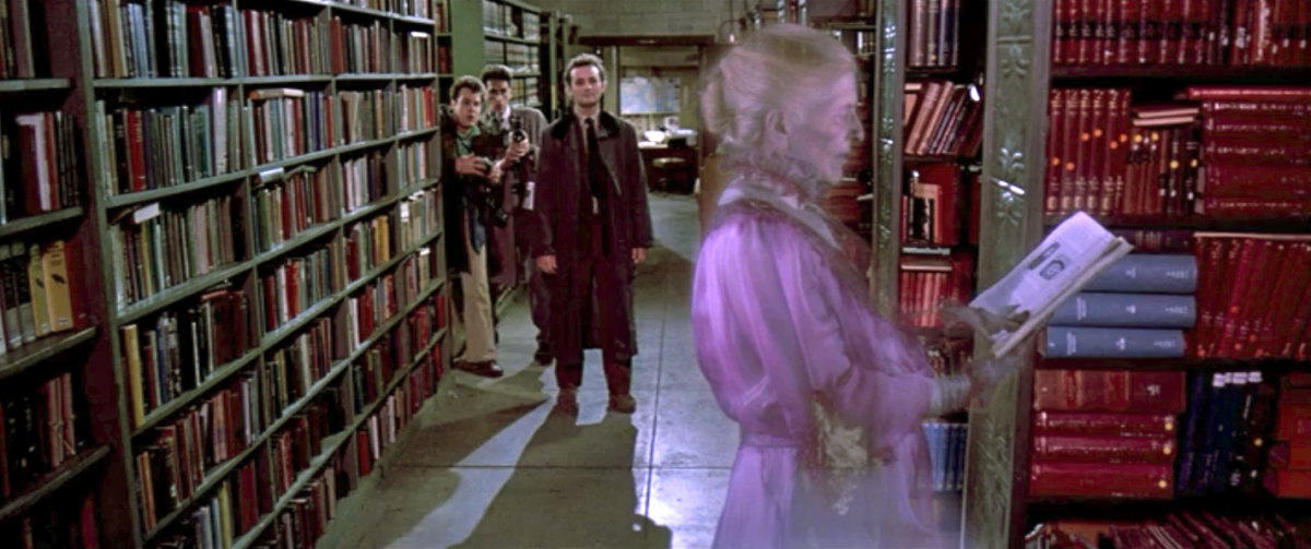 A ghost in a mauve Victorian dress reads a book in a public library, while the Ghostbusters watch.