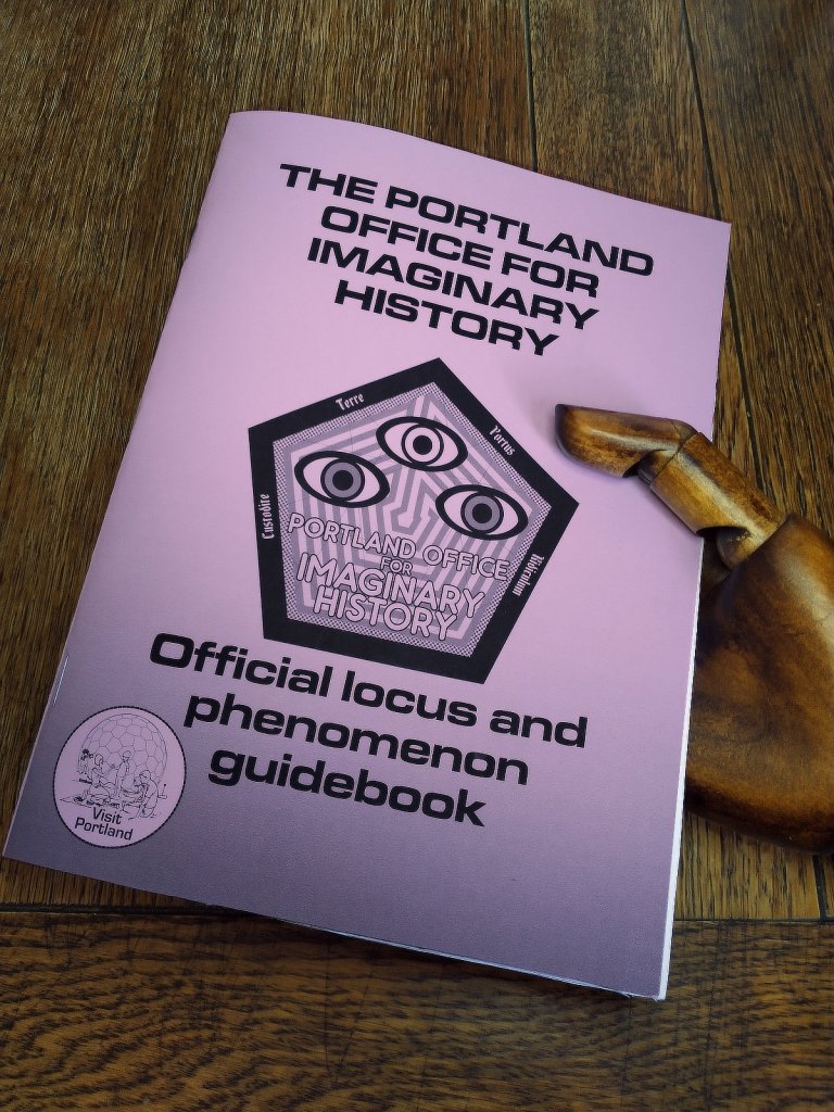 A wooden hand holding a Portland Office for Imaginary History Official Locus and Phenomenon Guidebook with a lilac cover.