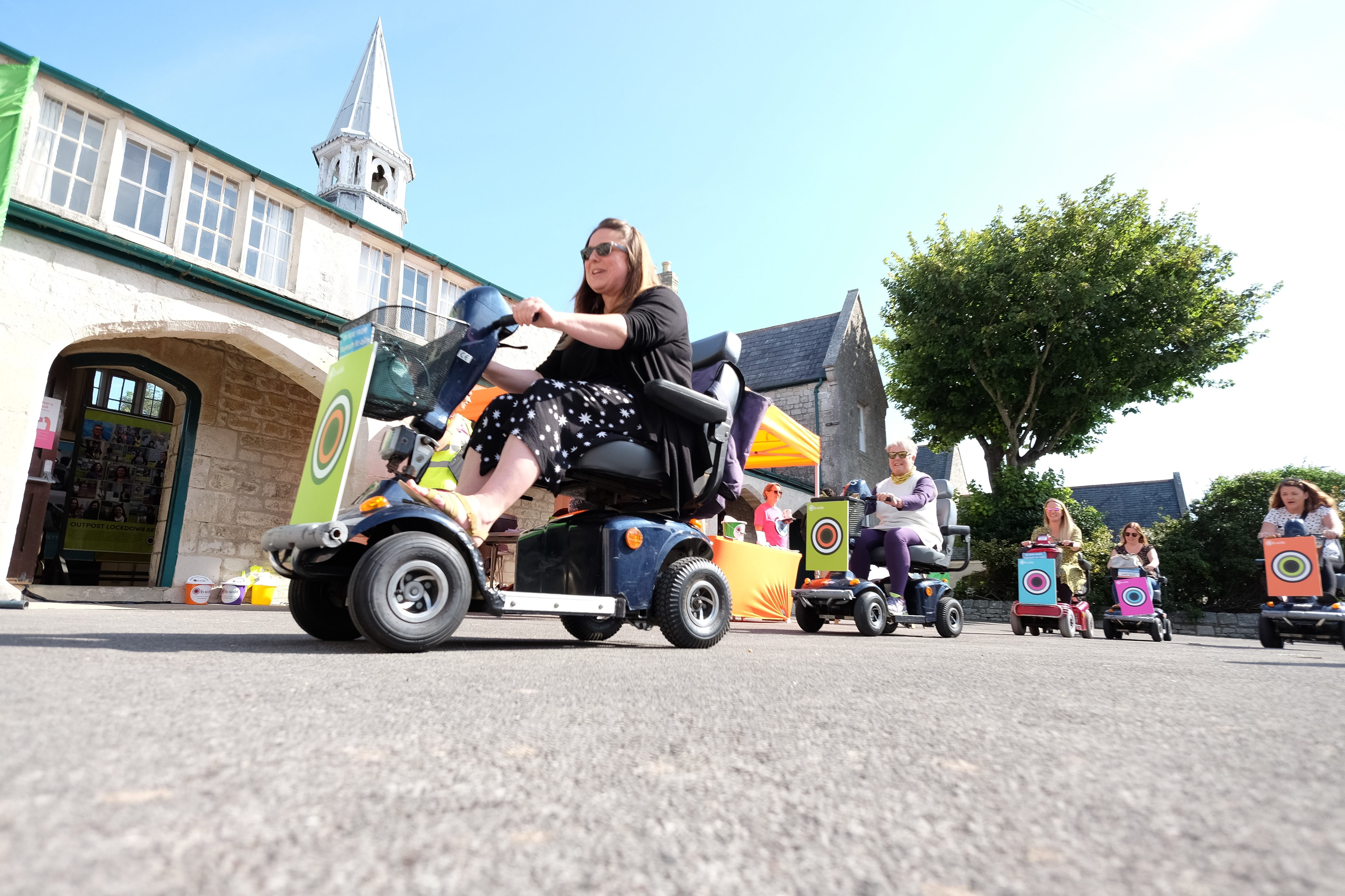 Seen from ground level, people on mobility scooters. Each scooter has a colourful sign on the front with a circular design and the the words "b-side festival".