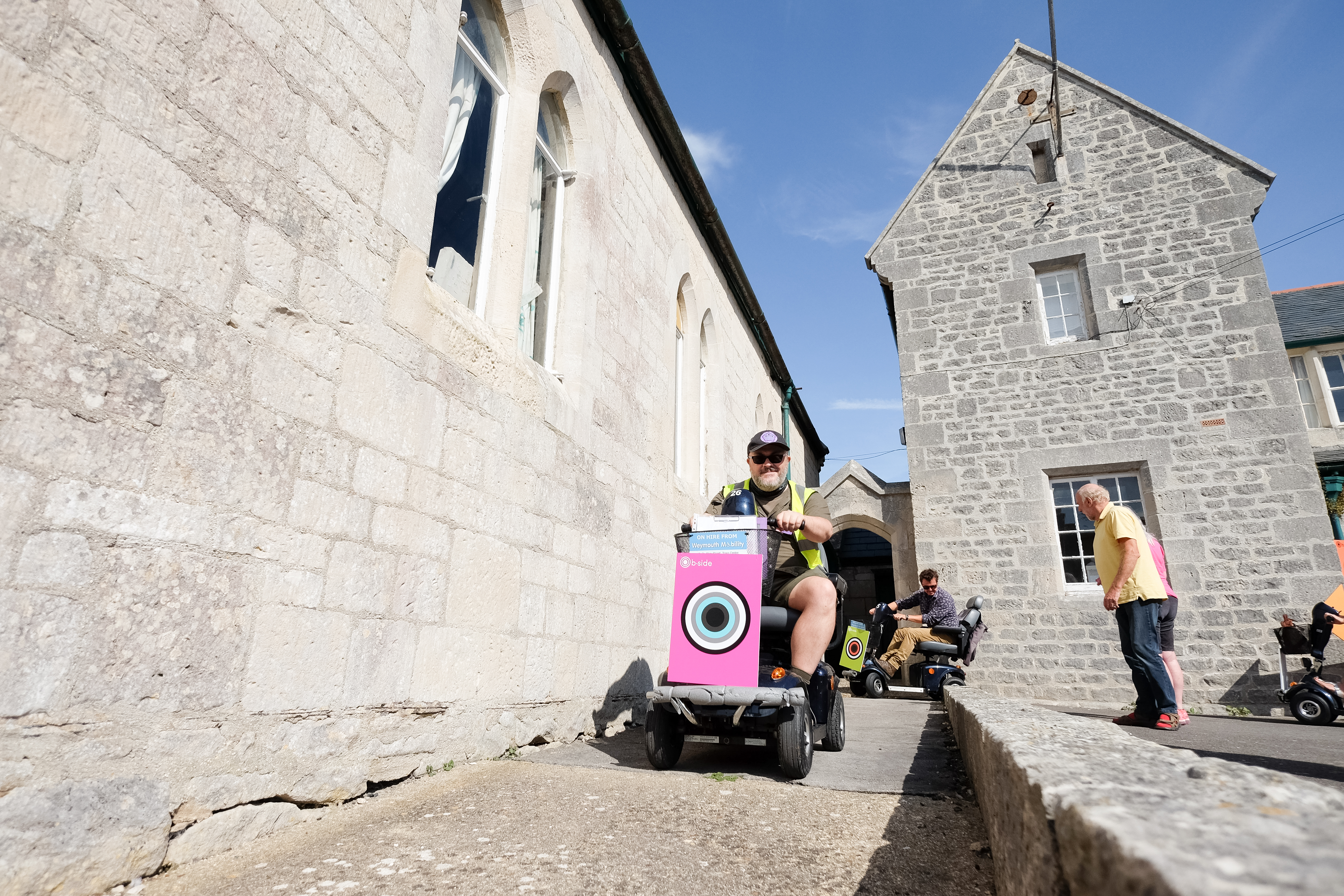 Alistair Gentry dressed as an Imaginary History ranger with a hi-vis jacket and cap, with mobility scooter users beside an old stone building.