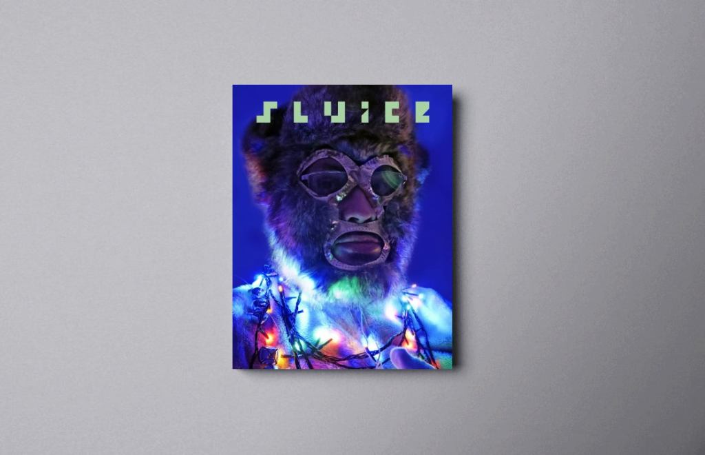 A vivid blue background and the title of the magazine "Sluice" at the top. The image is of a person wearing a brown and grey fur mask with gold-trimmed holes of the eyes, nose and mouth. The torso is dressed in white fur and wrapped in a combination of coloured Christmas lights and sleigh bells.