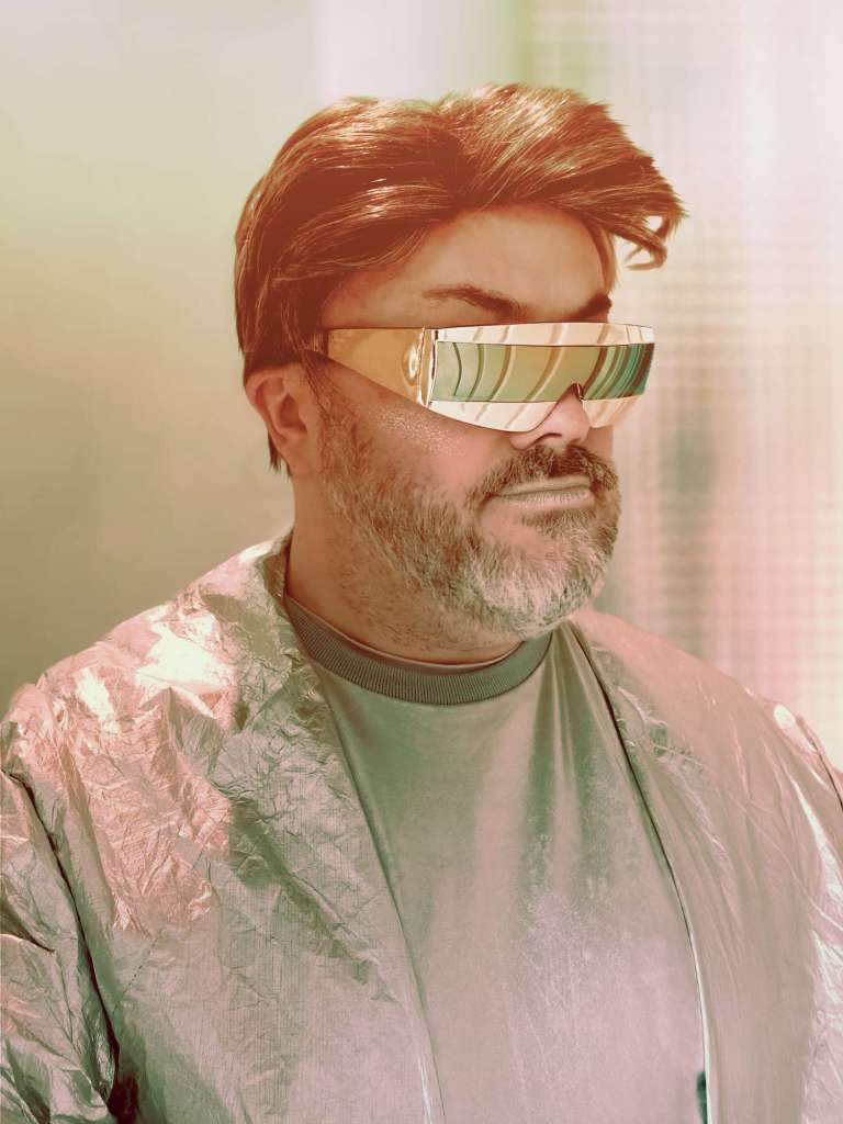 Alistair Gentry wearing a wig, gold mirrored wraparound glasses, silver lipstick, and a silver shirt under a silver coat.