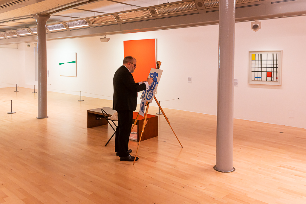 Wide shot of the artist Alistair Gentry, dressed in a black dinner suit and wearing dark glasses, working on a painting on a wooden easel. In the background is a white art gallery with a painting by Mondrian and several other paintings on it.