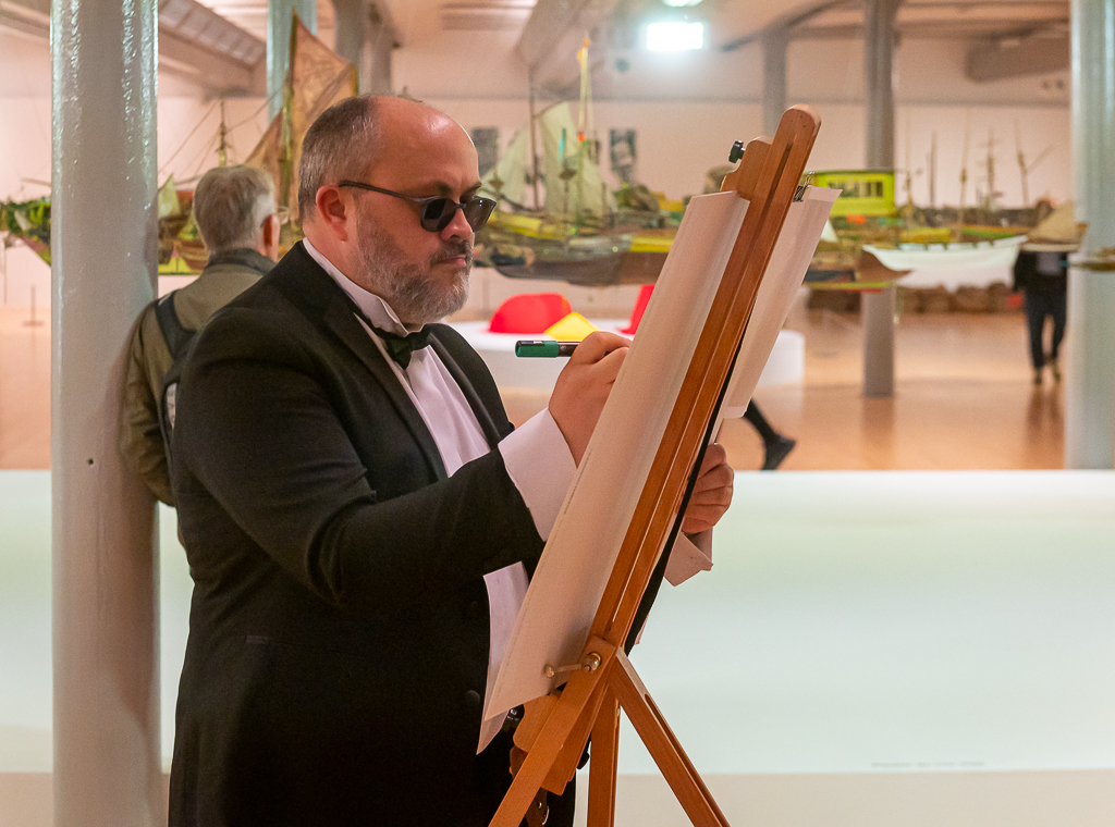The artist Alistair Gentry, dressed in a black dinner suit and wearing dark glasses, working on a painting on a wooden easel. In the background, out of focus, is an art gallery.