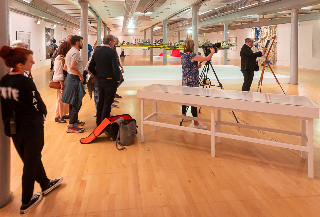 The artist Alistair Gentry is in the background, dressed in a black dinner suit and wearing dark glasses, working on a painting on a wooden easel. In the foreground, a person is filming with a camera on a tripod, and gallery visitors watch.