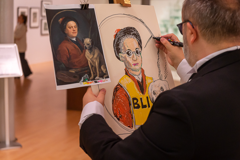 The artist Alistair Gentry, dressed in a black dinner suit and wearing dark glasses, working on a painting on a wooden easel. The painting is a copy of one by William Hogarth, who is now wearing a hi vis "blind" tabard and has a guide dog. In the background, out of focus, is an art gallery.