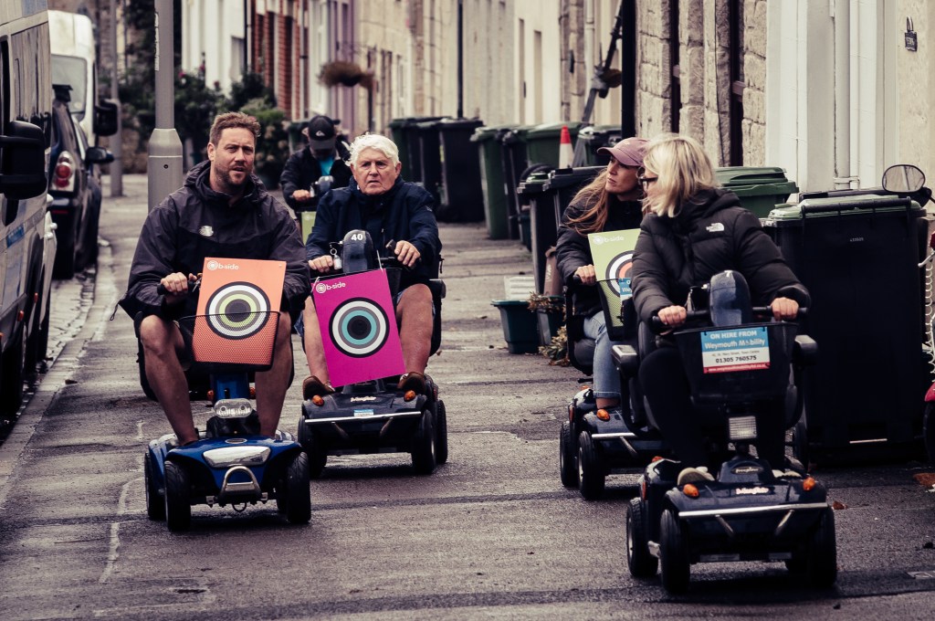 People in mobility scooters on a wide pavement, with terraced houses in the background.