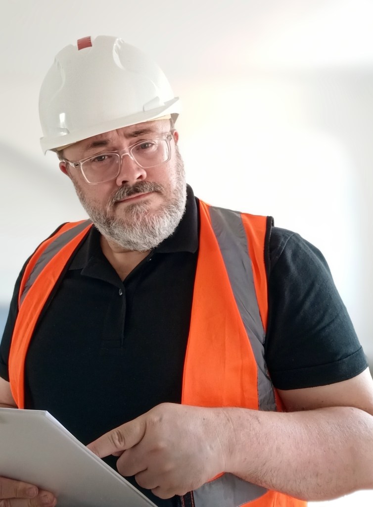 The artist Alistair Gentry, a white man with glasses and a grey beard, wearing a white hard hat, a black shirt, and an orange hi vis vest, pointing at a white clipboard.