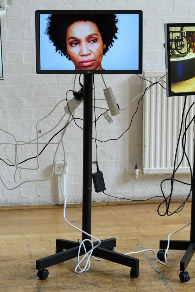 Installation view of Unbuilt Environments, photographed by Kirsten Holst. Closeup of one screen on its stand, showing a Black woman's face looking curiously to one side.