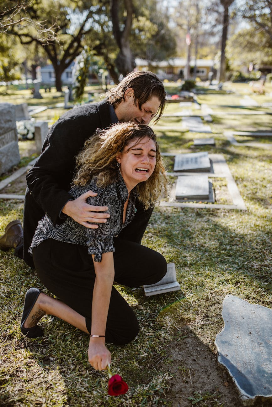 A stock photograph showing two people wearing black at a graveside in a well-kept cemetery in the daytime. A man is comforting a woman who is crying and holding a rose.